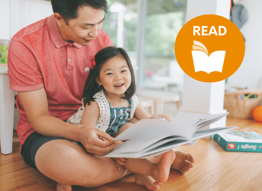 A young toddler sits on her father's lap reading a picturebook